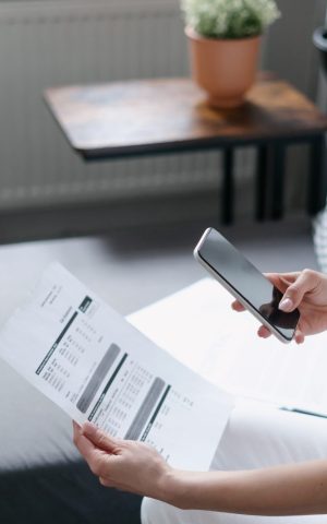 Une femme en tenue décontractée prenant en photo un document contenant des justificatifs de frais avec son smartphone, assise dans un intérieur moderne