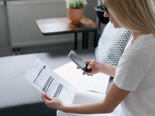 Une femme en tenue décontractée prenant en photo un document contenant des justificatifs de frais avec son smartphone, assise dans un intérieur moderne