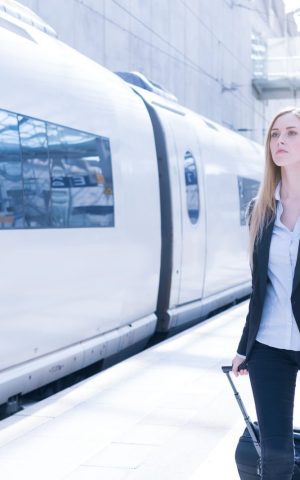 Une femme d'affaires en tenue professionnelle noire et blanche marchant sur un quai de gare à côté d'un train blanc moderne, tenant une valise à roulettes et un manteau.