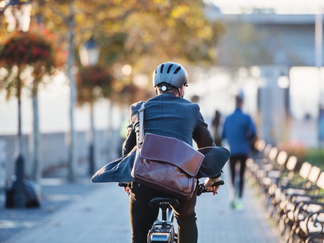 Un homme en costume portant un casque et un sac en cuir roule à vélo sur une promenade bordée d'arbres, par une journée ensoleillée d'automne.